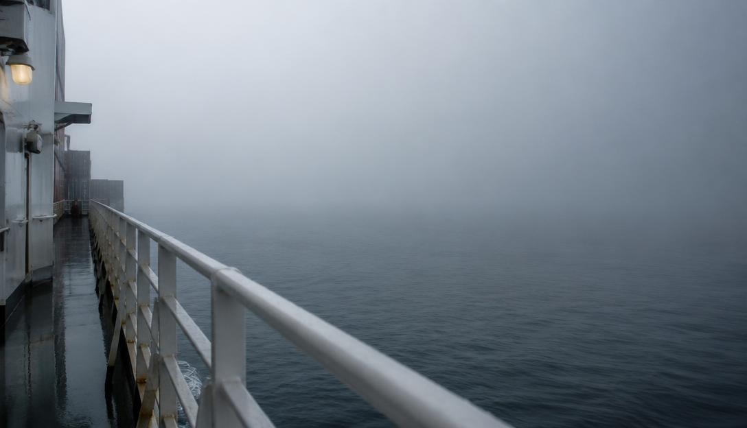 Wet container ship railings fading into dense silver fog on a calm ocean.